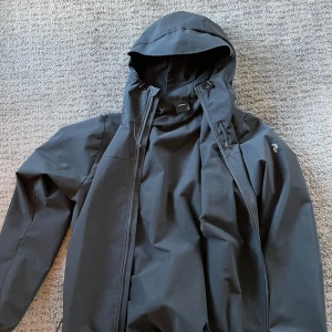 Svart vindjacka Peak Performance L - Svart vindjacka från Peak Performance med huva och dragkedja framtill. Jackan har minimalistisk design, diskret logga på bröstet och är tillverkad i ett slitstarkt syntetmaterial. Perfekt för blåsiga dagar och outdoor-aktiviteter. Säljes då den är för liten. Använd fåtal gånger