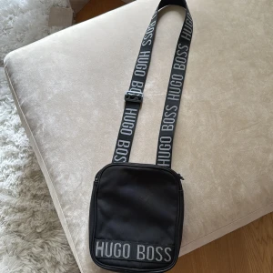 Hugo boss axelremsväska, svart  - Säljer nu min Hugo boss väska med justerbart band och dragkedja. Säljer nu för att jag har köpt en annan väska så den behövs inte längre, perfekt att lägga småsaker i (telefon nycklar osv) lätt att tvätta om det skulle komma smuts på och enkel att hantera. 