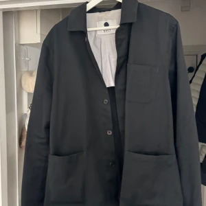 Svart overshirt från NN07 - Stilren svart overshirt från NN07 med klassisk krage, tre fickor framtill och knäppning med knappar. Perfekt att slänga över en t-shirt för en clean look. Materialet är bomull och modellen har en rak passform med lång ärm. Nypris 2399kr.