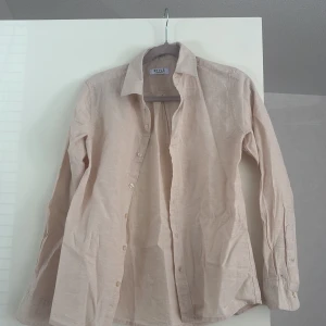 Beige linneskjorta från RYLS - Stilren beige skjorta från RYLS i svalt linnematerial. Klassisk krage, knäppning framtill och långa ärmar med manschett. Perfekt för dig som gillar en enkel och clean look med naturlig känsla.