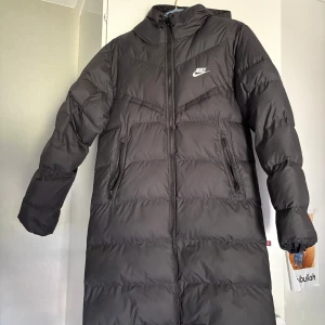 Svart lång dunjacka Nike Storm-FIT XS - Lång svart dunjacka från Nike med Storm-FIT-teknologi och Primaloft-foder. Jackan har huva, två dragkedjeförsedda fickor och hel dragkedja framtill. Perfekt för kalla dagar och riktigt snygg streetstil. Klassisk Nike-logga på bröstet.