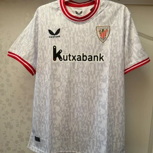 Athletic Club Bilbao vit matchtröja - Snygg vit Athletic Club Bilbao fotbollströja från Castore med subtilt mönster och röda detaljer på krage och ärmslut. Tryckta loggor och sponsor på bröstet och ryggen. Lätt och ventilerande material, perfekt för match eller träning.