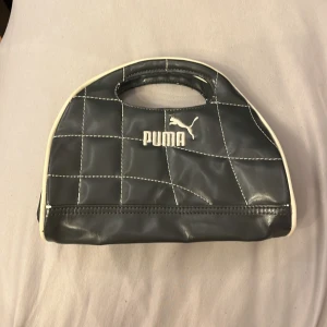 Svart handväska från Puma i syntet - Cool grå handväska från Puma i glansig syntet med vita detaljer och kontrastsömmar. Väskan har ett stilrent broderat Puma-logo framtill, praktiskt bärhandtag och dragkedja upptill. Perfekt storlek för det viktigaste och ger en sportig vibe till din outfit. Kan även användas som sminkväska :)