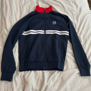 Sergio Tacchini blå track jacket M - Snygg blå track jacket från Sergio Tacchini med röd krage och vita dubbla ränder över bröstet. Jackan har dragkedja framtill, två fickor och logga på bröstet. Perfekt för en sportig vibe. Köpt för 899 kr säljer den för att endast 350 nästan helt oanvänd
