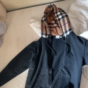 Svart Burberry hoodie med rutigt foder - Svart hoodie från Burberry med dragkedja och klassiskt rutigt foder i huvan. Tröjan har snörning vid huvan och två fickor framtill. Perfekt för dig som vill ha en stilren och lyxig streetwear-look.