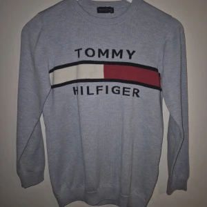 Ljusblå stickad Tommy Hilfiger tröja, storlek 176 - Snygg ljusblå stickad tröja från Tommy Hilfiger med klassisk logga och ränder i vitt, rött och svart över bröstet. Tröjan har rund halsringning och långa ärmar, perfekt för dig som gillar stilren och sportig look. Storlek 176 cl                                                                   Fint skick, dock ett pyttelitet som jag upptäckte när jag granskade den. Såg ut som ett dammkorn så är litet. Djur och rökfritt hem 