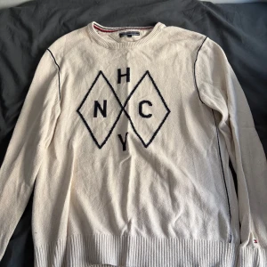 Vit stickad tröja Tommy Hilfiger L - Vit/crämig vit stickad tröja från Tommy Hilfiger i storlek L. Tröjan har svarta detaljer, ribbade muddar och ett stort grafiskt mönster med bokstäver på bröstet. Klassisk rund halsringning och diskret logga vid nederkanten. Perfekt för dig som gillar stilrena plagg med en twist.