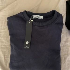Mörkblå sweatshirt från Stone Island - Snygg mörkblå sweatshirt från Stone Island med klassisk rund halsringning och ribbade muddar. Tröjan är i mjuk bomull och har en clean look utan tryck eller mönster. Perfekt för dig som gillar stilrena och exklusiva plagg. Loggan som sätts dit på tröjan säljs tyvärr inte med den.