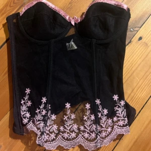 Svart korsett med rosa spets Wild Designs - Supersnygg svart korsettopp från Wild Designs med rosa satin-detaljer upptill och broderade blommor i rosa längs nederkanten. Perfekt för att sticka ut med coola detaljer och blommig spets.