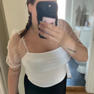 Vit topp med puffärmar och prickigt nät - Superfin vit topp med transparenta puffärmar i prickigt nät. Toppen har en rak, croppad passform och fyrkantig urringning. Ärmarna är korta och ger en romantisk vibe. Perfekt att styla med jeans eller kjol för en trendig look.
