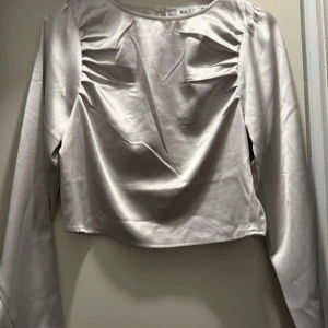 Ljus champagnefärgad blus från NA-KD - Säljer en elegant ljus champagnefärgad blus från NA-KD med långa ärmar och rynkade detaljer vid axlarna. Blusen har en mjuk, glansig satinfinish och rund halsringning. Perfekt för dig som gillar stilrena och trendiga plagg.