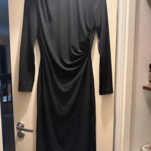 Svart långklänning från Vila - Svart långklänning från Vila med lång ärm och snygg drapering i midjan. Klänningen har en stilren och enkel design som ger en elegant siluett. Tillverkad i ett mjukt och följsamt material som känns bekvämt mot huden.