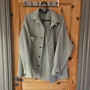 Ljusgrå manchester overshirt - Snygg ljusgrå overshirt i manchester med klassisk krage, stora knappar och bröstficka. Jackan har en loose passform och långa ärmar med knapp vid ärmslut. Perfekt att slänga över en hoodie eller t-shirt för en chill look.