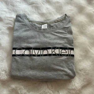 Grå Calvin Klein tröja med logga - Snygg grå tröja från Calvin Klein med stor logga tryckt över bröstet i svart och vitt. Rund halsringning och mjukt bomullsmaterial som är skönt mot huden. Perfekt för chill dagar eller när du vill ha en clean look.
