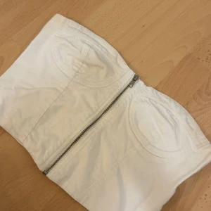 Vit korsett-tubtopp  - Snygg vit korsett-tubtopp från Gina Tricot med markerade kupor och dragkedja framtill. Toppen är axelbandslös och har en stilren, croppad passform. Perfekt att styla med jeans eller kjol. Köpt för 299kr och har aldrig använt den då den är för stor för mig. (kommer strykas innan den skickas) pris kan diskuteras.