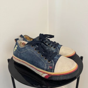 Blå denim sneakers från Rooster - Säljer ett par unika sneakers från Rooster i blått denimtyg med vita mockadetaljer på tån och coola röda och svarta detaljer på sulan. Skorna har snörning, blommigt foder och en liten döskalle på sidan. Perfekt för dig som gillar streetstyle och vill sticka ut.
