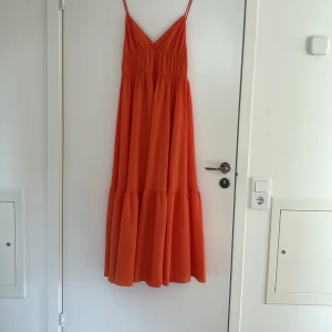Orange långklänning  - Snygg orange långklänning från H&M i storlek 38. Klänningen har smala axelband, v-ringad urringning och ett luftigt, voluminöst fall. Perfekt för varma dagar och sommarhäng.