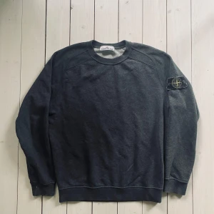 Mörkgrå sweatshirt från Stone Island - Säljer en mörkgrå sweatshirt från Stone Island med klassisk rund hals och ribbade muddar. Tröjan har den ikoniska Stone Island-patchen på vänster ärm och är tillverkad i mjuk bomull. Perfekt för dig som gillar streetwear och snygga detaljer.