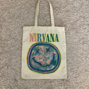 Tygväska - Säljer en beige totebag med Nirvana-tryck på framsidan. Perfekt för att ha dator eller annat i. Obs: Nirvana är ett band, inte ett märke som många tror. Läs helst min bio innan du köper💗