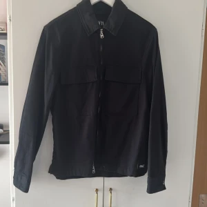 Svart overshirt från Zara - Svart overshirt med klassisk krage och två stora bröstfickor med lock. Jackan har hel dragkedja framtill och långa ärmar med knapp vid ärmslut. Materialet är slätt och känns lätt, perfekt för lager-på-lager. Enkel och stilren design som funkar till många outfits.