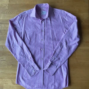 Randig lila skjorta från Stenströms - Snygg lila skjorta från Stenströms i slimline-modell, tillverkad i tvålagers superbomull. Skjortan har vita ränder, klassisk krage och knappar framtill. Långärmad med manschettknappar och stilren passform. Perfekt för dig som gillar färg och mönster. Bortsett från hålet är den sparsamt använd.