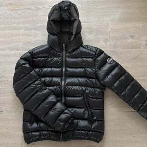 Svart Colmar pufferjacka med huva - Säljer en svart pufferjacka från Colmar Originals med huva och snygga breda kanaler. Jackan har dragkedja framtill, två sidofickor med dragkedja och Colmar-logga på ärmen. Materialet är glansigt och jackan är fodrad med dun för extra värme. Perfekt för kalla dagar. Högra fickans dragkedja har fastnat och går inte att öppna, ev kan man fixa det hos en skräddare.