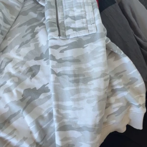 Alpha Industries camo bomberjacka - Snygg bomberjacka från Alpha Industries i ljusgrått och vitt camouflagemönster. Jackan har ribbade muddar, en ficka med dragkedja på ärmen och orange foder på insidan. Perfekt för dig som gillar streetwear och vill sticka ut.