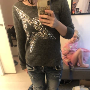 Mörkgrå stickad tröja med paljettstjärna - Mysig mörkgrå stickad tröja med lång ärm och en stor glittrig paljettstjärna framtill. Tröjan har en offshoulder-look och sitter tight. Perfekt för dig som gillar lite extra bling i vardagen. Y2k vibbar