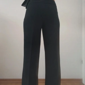 Svarta vida kostym byxor med veck  - Snygga svarta byxor med hög midja och wide fit. De har raka, vida ben och är perfekta för en clean och trendig look. Materialet känns mjukt och följsamt, vilket gör dem riktigt sköna att bära. Passar dig som gillar stilrena och enkla plagg.Mycket bra skick!  Storlek M/38  Högmidjade.  Totallängd ca 100 cm.  De passar M/L, skicka meddelande för mer exakta mått.   Skickas samma dag eller nästa dag.  För exakta mått eller andra frågor skicka ett meddelande.
