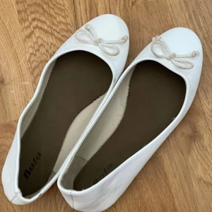 Vita ballerinaskor med rosett från Bata - Säljer ett par vita ballerinaskor från Bata med rund tå och söt rosettdetalj framtill. Skorna har en platt sula och är gjorda i ett syntetmaterial som känns mjukt och bekvämt. Perfekt för dig som gillar enkel och stilren design med en feminin touch.