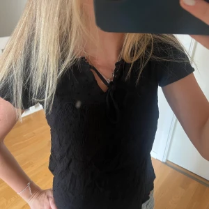 Svart blus med knyt från JDY - Svart blus från JDY med korta ärmar och snygg knytning vid halsen. Blusen har diskreta prickar i tyget och volangdetaljer runt halsringningen. Jättefin i jättebra skick!!❤️