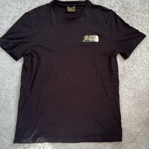 Svart EA7 t-shirt med guldlogga äkta  - Svart t-shirt från EA7 Emporio Armani med en snygg guldig logga på bröstet. Klassisk rund hals och korta ärmar. Materialet är mjuk bomull som känns skönt mot huden. Perfekt för dig som gillar stilrena och exklusiva plagg med en touch av lyx.
