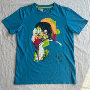 Blå Messi t-shirt från Adidas - Snygg blå t-shirt från Adidas med Messi-tryck på framsidan i färgglada toner som limegrönt, rött och orange. T-shirten har korta ärmar, rund hals och är gjord i bomull med Climalite-teknologi. Består även av polyester. Perfekt för dig som gillar fotboll och coola prints. Den är använd men ända slitaget är på etiketten i nacken som är lite sliten, annars i bra skick, hel och ren. Både trycket på magen och Messi-loggan är i väldigt fint skick.