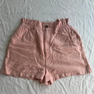 Rosa linneshorts från Bik Bok - Snygga rosa shorts från Bik Bok i storlek XS. Tillverkade i linne och viskos med framfickor. Modellen är högmidjad med rynkad midja, bälteshällor och knappstängning samt dragkedja framtill. Ingår ett matchande ljusrosa brett snöre. Perfekta för varma dagar. Använda ett fåtal gånger.
