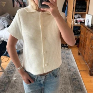 Vit stickad kofta med kort ärm - Säljer en vit stickad kofta med korta ärmar och klassisk krage. Koftan har en rak passform och stängs med matchande knappar framtill. Perfekt att styla med jeans eller kjol för en clean look. Inte min bild💗