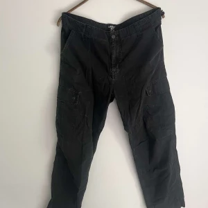 Svarta cargopants från Carhartt WIP - Svarta cargopants från Carhartt WIP med rutigt mönster i tyget och flera praktiska fickor på sidorna och bak. Byxorna har normal passform och är tillverkade i slitstark bomull. Klassisk Carhartt-logga på benet och knappstängning framtill. Gott skick förutom liten reva på ena benet. Nypris 1199kr