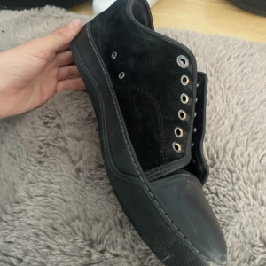 Svarta sneakers med mockadetaljer - Svarta sneakers med snygg kombination av mocka på sidorna och slät tå i syntet. Skon har rund tå, platt sula och metallöglor för snörning. Perfekt för dig som gillar stilrena och klassiska skor med lite edge.