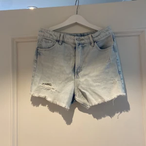 Ljusblå jeansshorts från H&M - Säljer ett par ljusblå jeansshorts från H&M med hög midja och relaxed passform. Shortsen har slitna detaljer, fransig kant nedtill och klassiska fem fickor. Perfekt för dig som gillar en chill och trendig stil.