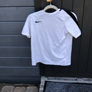 Nike Dri-Fit vit t-shirt L - Vit Nike Dri-Fit t-shirt med svart logga på bröstet och svarta mesh-detaljer över axlarna. Materialet är lätt och andas, perfekt för sport och träning. Rund hals och korta ärmar, med ventilerande panel på ryggen för extra komfort.