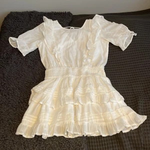Vit volangklänning från Gina Tricot - Supersöt vit kortklänning från Gina Tricot med volanger och smock i midjan. Klänningen har korta ärmar, rund hals och flera lager med volanger på kjolen. Materialet är lätt och ger en luftig känsla, perfekt för sommarens alla tillfällen. Säljes då den inte längre används!