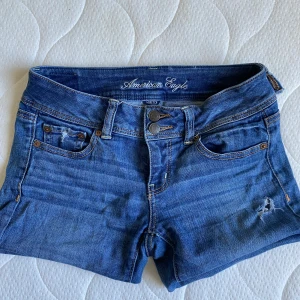 Lågmidjade jeansshorts American Eagle - Snygga blå jeansshorts från American Eagle med låg midja och kort passform. dubbla knappar, rå kant nedtill. Väldigt Stretchigt denimmaterial som sitter skönt och formar sig efter kroppen. 📏mått: ca 38-39 cm rakt över
