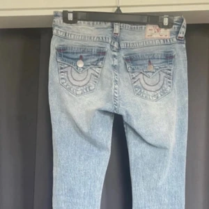 True Religion ljusblå bootcut jeans - Säljer ett par ljusblå jeans från True Religion med bootcut passform. Jeansen har slitna detaljer på framsidan, klassiska fickor med dekorativa sömmar och silverfärgade knappar. Materialet är jeans med snygg tvätt och röda kontrastsömmar på bakfickorna.