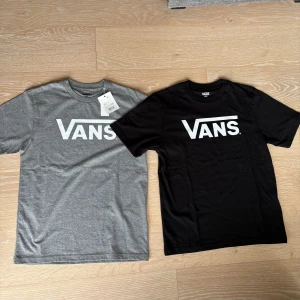 Två Vans t-shirt svart och grå, JR Large - Två klassiska Vans t-shirts i storlek JR Large, en i svart och en i grått. Helt oanvända en med prislapp kvar och en utan. Båda har den ikoniska Vans-loggan tryckt i vitt framtill med en rund hals och korta ärmar. Pris för båda två, säljs endast ihop.                                     Motagare står för frakt.