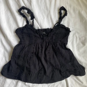 Svart linnetopp med spetsdetaljer - Supersöt svart linnetopp från Bikbok  med volangkant och smock baktill. Axelbanden är rynkade och toppen har fina spetsdetaljer längs hela framsidan. Luftig och flowy passform som ger en chill vibe. Perfekt att matcha med jeans eller kjol.