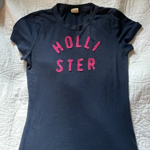Svart Hollister t-shirt med rosa tryck - Svart t-shirt från Hollister med korta ärmar och rund hals. Framsidan har ett stort rosa Hollister-tryck i broderad stil. Modellen är figurnära och gjord i mjuk bomull, perfekt för en chill och stilren look.