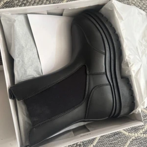 Svarta chelsea boots med grov sula - Säljer ett par svarta chelsea boots med chunky, mönstrad sula och elastiska paneler på sidorna. Bootsens ovandel är i slätt syntetmaterial och har en rund tå. Perfekt för dig som gillar stilrena och robusta skor med edge.