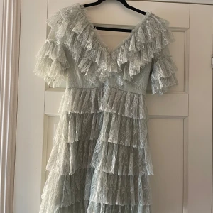 Ljus grön spetsklänning med volanger By Malina - Supersöt ljus spetsklänning från By Malina med flera lager volanger och v-ringad hals. Klänningen har korta ärmar och är tillverkad i spets med sidenfoder