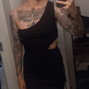 Svart oneshoulder kortklänning - Säljer en svart kortklänning med oneshoulder-design och snygg cut-out på sidan. Klänningen har en tight passform och cool snörningsdetalj i midjan. Perfekt för dig som vill sticka ut med en edgy och modern look.