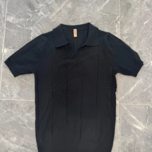 Svart pikétröja med krage - Svart pikétröja från Cos med klassisk krage och v-ringad hals. Tröjan har korta ärmar och är tillverkad i mjukt bomullsmaterial. Snygga vertikala sömmar framtill ger en stilren look. Perfekt för dig som gillar minimalistisk stil.