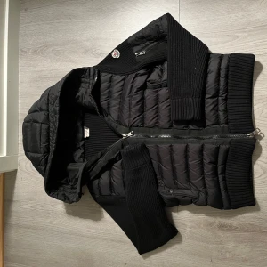 Svart dunväst med huva från Moncler - Snygg svart dunväst från Moncler med stickade partier på sidor, ärmar och rygg. Västen har en quiltad framsida, huva och dragkedja framtill. Ikonisk Moncler-logga på axeln. Perfekt för lager-på-lager och streetstyle. Nsf finns. Kan ej garantera äktheten 
